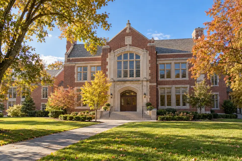 Red-brick school building with a stone arched entrance, framed by autumn trees and a sunlit lawn