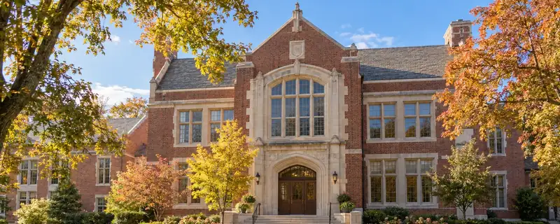 Red-brick school building with a stone arched entrance, framed by autumn trees and a sunlit lawn